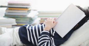 Child covering his head with a notebook while sitting beside other piles of notebooks.