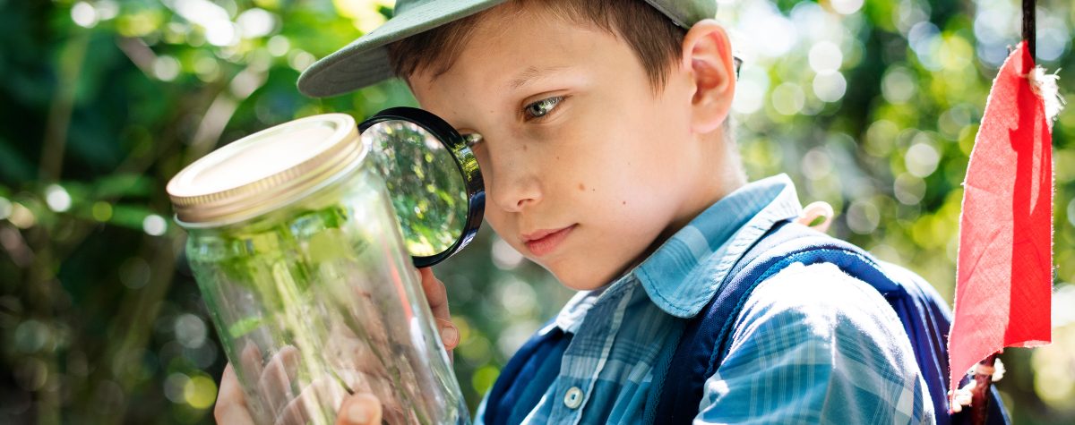 A young boy stands outdoors wearing a blue plaid shirt, green hat, and blue backpack. He holds a magnifying glass and examines a plant inside a jar.