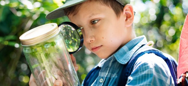 A young boy stands outdoors wearing a blue plaid shirt, green hat, and blue backpack. He holds a magnifying glass and examines a plant inside a jar.