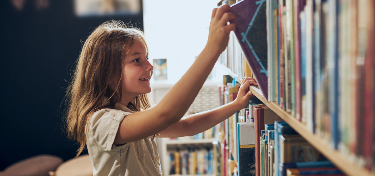 A young girl with long brown hair smiles as she reaches up to pull a book from a library shelf.