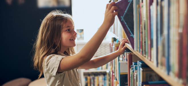 A young girl with long brown hair smiles as she reaches up to pull a book from a library shelf.