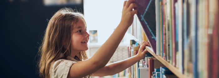 A young girl with long brown hair smiles as she reaches up to pull a book from a library shelf.