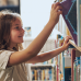 A young girl with long brown hair smiles as she reaches up to pull a book from a library shelf.