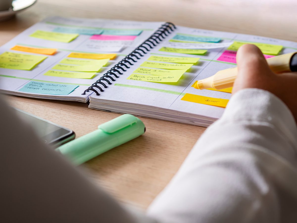 Close-up of agenda organize with color-coding sticky notes for time management. Productive schedule for appointments and reminders. Hand holding a yellow highlighter marker. Organization and planning