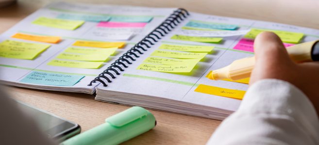 Close-up of agenda organize with color-coding sticky notes for time management. Productive schedule for appointments and reminders. Hand holding a yellow highlighter marker. Organization and planning