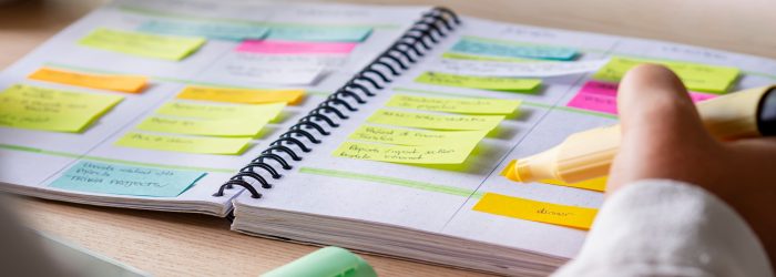 Close-up of agenda organize with color-coding sticky notes for time management. Productive schedule for appointments and reminders. Hand holding a yellow highlighter marker. Organization and planning